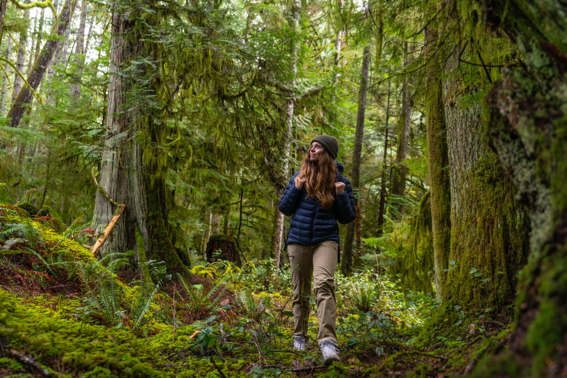 A woman with brown hair, a toque and a backpack walks through a lush green forest in British Columbia.